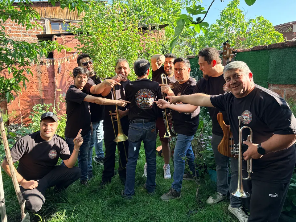 Los músicos de La Banda del Papi con poleras negras, riendo y apuntando a la cámara en un jardín